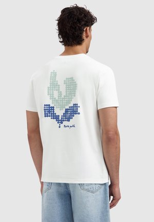 Man draagt een wit T-shirt met een abstract patroon van blauwe en grijze stippen en de tekst "Pure path" op de achterkant, gecombineerd met lichtblauwe spijkerbroek.