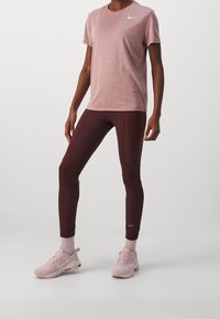 Osoba ubrana w jasnoróżową koszulkę Nike, ciemnoróżowe legginsy, różowe skarpetki oraz różowe buty Nike stojąca na jednolitym tle.