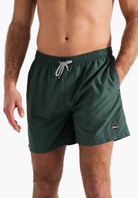 Shorts de bain vert foncé en tissu léger, avec une taille élastique dotée d'un cordon de serrage et un patch logo sur le côté.
