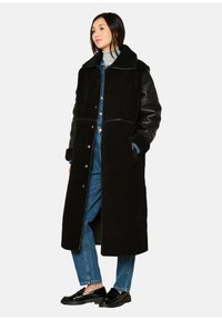 Zwarte jas met een wollenachtige textuur en leren mouwen, met een brede kraag en drukknoopsluiting, gedragen over denim.