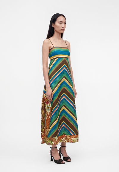 Γυναίκα με αμάνικο, μακρύ φόρεμα μέχρι τους αστραγάλους με πολύχρωμες διαγώνιες λωρίδες και λουλουδάτα σχέδια, συνδυασμένο με μαύρα ψηλοτάκουνα σανδάλια.