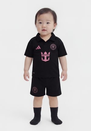 Niño pequeño con cabello corto, vestido con ropa deportiva negra de Adidas y calcetines, de pie contra un fondo blanco liso.
