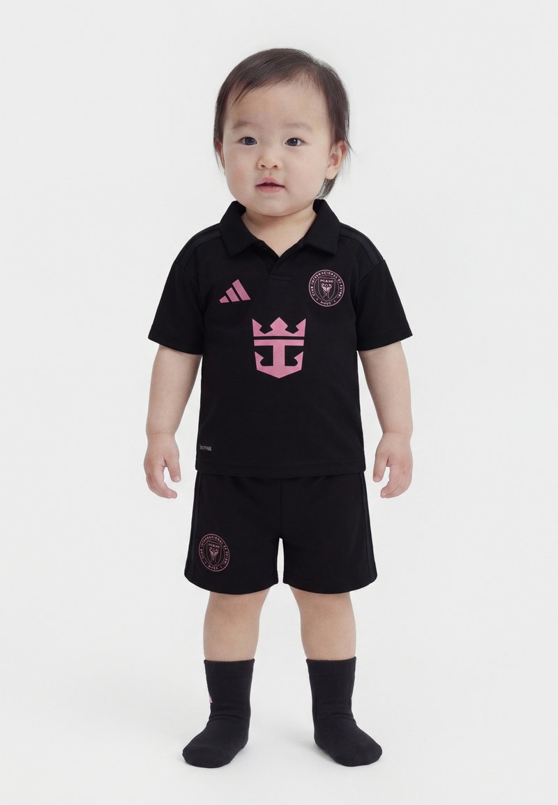 Παιδάκι με κοντά μαλλιά που φοράει μαύρη αθλητική φόρμα Adidas και κάλτσες, στέκεται μπροστά σε απλό λευκό φόντο.