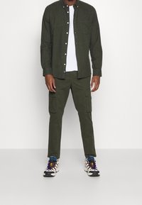 Man med mörkgröna cargobyxor och matchande skjorta över en vit T-shirt, samt vita, blå och lila sneakers.