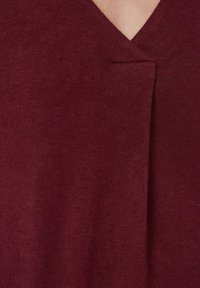 Tuvo plāna skats uz bordo V veida izgriezumu ar salocītu plisējumu tuvumā pie kakla izgriezuma.