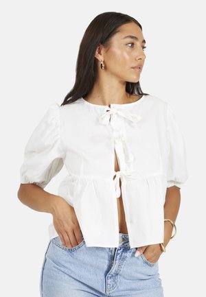 Kobieta w białej bluzce z bufkami i kokardami z przodu oraz jasnoniebieskich dżinsach, stojąca bokiem z jedną ręką w kieszeni.