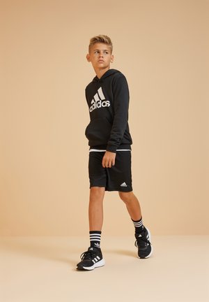 Felpa nera con logo bianco Adidas, pantaloni corti neri, scarpe nere con dettagli bianchi e calzini a righe bianche e nere. Design minimalista.