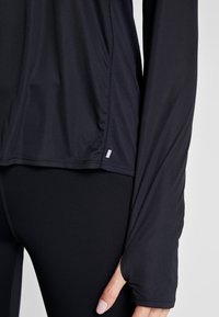 Lähikuva henkilöstä, joka käyttää mustaa pitkähihasta urheiluversoa, jossa on peukalonaukko, sekä mustia legginssejä. Käsi lepää sivulla valkoista taustaa vasten.