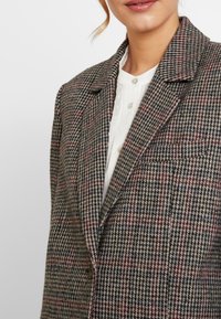 Sakó s kostkovaným vzorem houndstooth v hnědých, béžových a červených tónech. Obsahuje středový límec, zapínání na jeden knoflík a strukturovanou tkaninu.