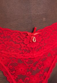 Lingerie in pizzo rosso con motivi floreali, caratterizzata da un piccolo fiocco decorativo e un logo in oro. La texture è trasparente e intricata.