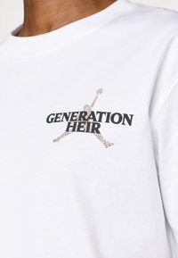 Balta medvilninė marškinėliai su juodu užrašu "GENERATION HEIR" ir auksiniu Jumpman logotipu kairėje krūtinės pusėje. Trumpomis rankovėmis, apvaliu kaklu.