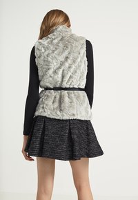 Ljust grå fuskpälsväst med en texturerad yta och ett svart bälte, bärs över en svart långärmad topp och en svart utsvängd kjol.