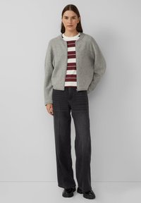 Graue, gestrickte Jacke mit Rundhalsausschnitt und Reißverschluss, getragen über einem weißen, gestreiften T-Shirt. Kombination mit hoch taillierten, dunklen Jeans.