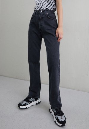 Persona que lleva vaqueros negros de corte recto, zapatillas negras y blancas, y una camiseta de rayas en blanco y negro, de pie sobre un suelo gris.