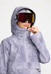Chaqueta exterior de color púrpura claro con capucha, que presenta un cierre medio con cremallera y un tejido texturizado y resistente al agua. Gafas de esquí negras y amarillas.