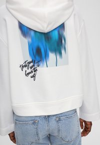 Weißer Hoodie mit einem blauen abstrakten Design auf der Rückseite und handgeschriebenem Text "vertrau deiner Seele, sie kennt den Weg" in dunkler Schrift.