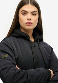Veste bomber noire avec une fermeture éclair à l'avant, dotée d'une capuche noire et d'un patch vert sur la manche gauche. Tissu texturé, design ajusté.