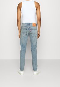 Man som bär ljusblå denimjeans, vit linnetröja och vita sneakers står vänd bort från kameran mot en enfärgad bakgrund.