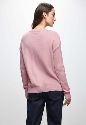 Donna con lunghi capelli scuri, che indossa un maglione rosa chiaro e jeans blu scuri, girata leggermente di lato su uno sfondo chiaro e semplice.
