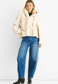 Woman with blonde hair wearing cream fur jacket, blue jeans, black ankle boots, standing against white background with one hand in pocket.