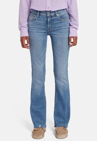 Jean flare bleu clair avec une taille mi-haute, design classique à cinq poches, décoloration subtile et ourlet effiloché. Porté avec des chaussures beiges.