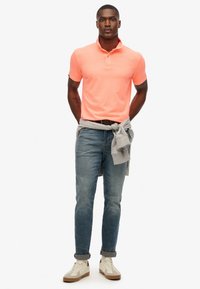 Lichtoranje polo met kraag, gecombineerd met blauwe jeans en witte sneakers. Een grijze sweatshirt is om de taille geknoopt.