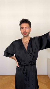 Črn satenski kimono z širokimi rokavi in vezanim pasom. Gladka tekstura, do kolen, z V-izrezom. Minimalističen dizajn na svetli podlagi.