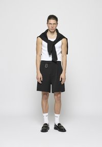 Shorts noirs avec des plis, débardeur blanc sans manches, et un pull noir drapé sur les épaules. Des mocassins noirs et des chaussettes blanches à mi-mollet complètent le look.