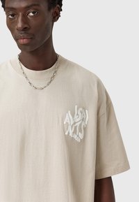 Man wearing beige t-shirt with white distorted text "AllSaints London" on left chest and silver chain necklace, neutral background.