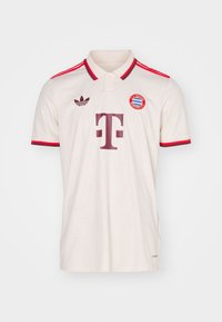 Valkoinen FC Bayern München -jalkapalloilupaita, jossa on punaiset raidat hartioilla ja kauluksessa, Adidas-logo ja Telekomin sponsorilogo edessä.