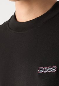 Gros plan d’une personne portant un t-shirt noir à col rond avec le mot BOSS brodé en rose et blanc sur la poitrine.