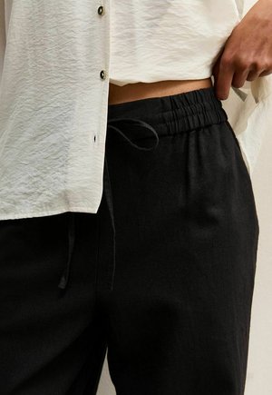 Persoon die zwarte elastische taillebroek verstelt, een wit overhemd met knopen draagt dat gedeeltelijk omhoog is getild, waardoor een blote buik zichtbaar is.