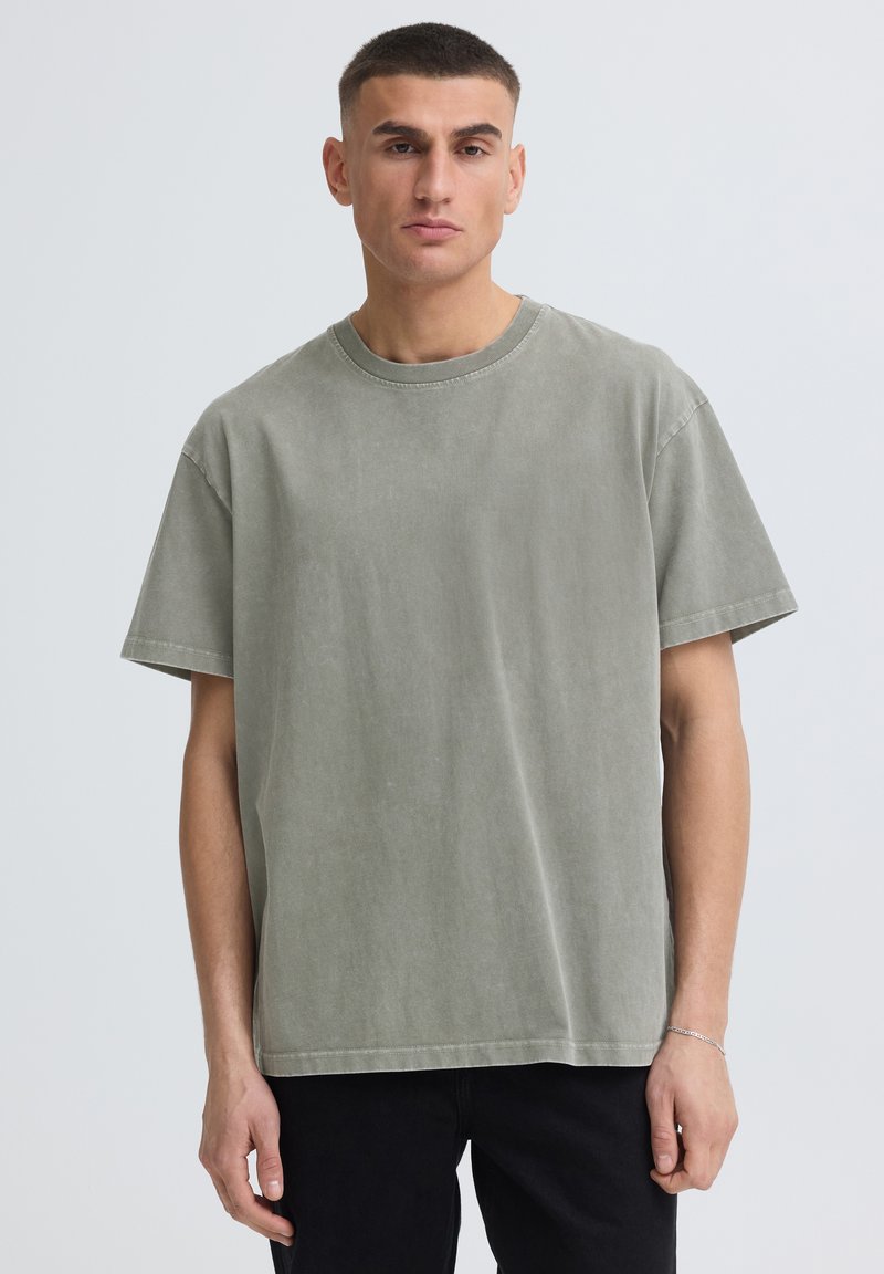 Homme aux cheveux courts portant un t-shirt vert délavé oversize et un pantalon noir, debout devant un fond clair uni.