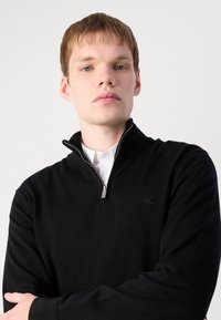 Melna adīta džemperis ar rāvējslēdzēja apkakli un smalku logo detaļu. Iezīmē gludu tekstūru un standarta piegriezumu, uzlikts uz baltas krekla.