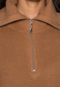 Gros plan sur un pull en tricot côtelé marron avec une fermeture éclair en métal partiellement ouverte au col sur une couche de col roulé haut.