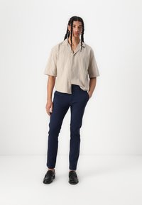 Chemise beige à manches courtes, boutonnée, avec des rayures verticales, associée à un pantalon slim-fit bleu marine et des mocassins noirs. Texture lisse sur toute la surface.