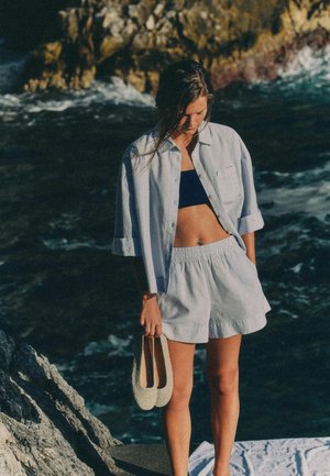Young woman standing on rocky shore wearing light shirt, shorts, holding beige shoes, looking down toward ocean waves.