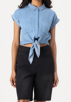 Mujer con una camisa vaquera de manga corta de color azul claro anudada en la cintura y pantalones cortos vaqueros negros de talle alto.