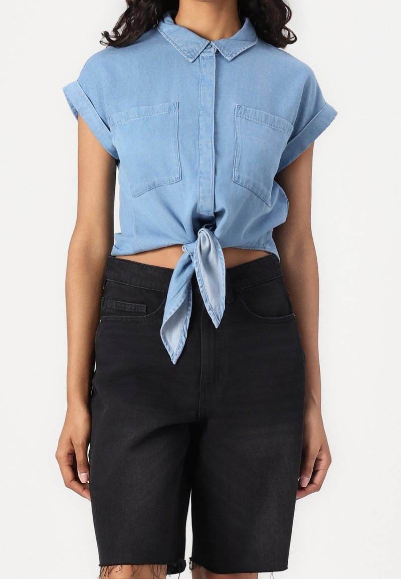Femme portant une chemise en jean bleu clair à manches courtes nouée à la taille avec un short en jean noir taille haute.