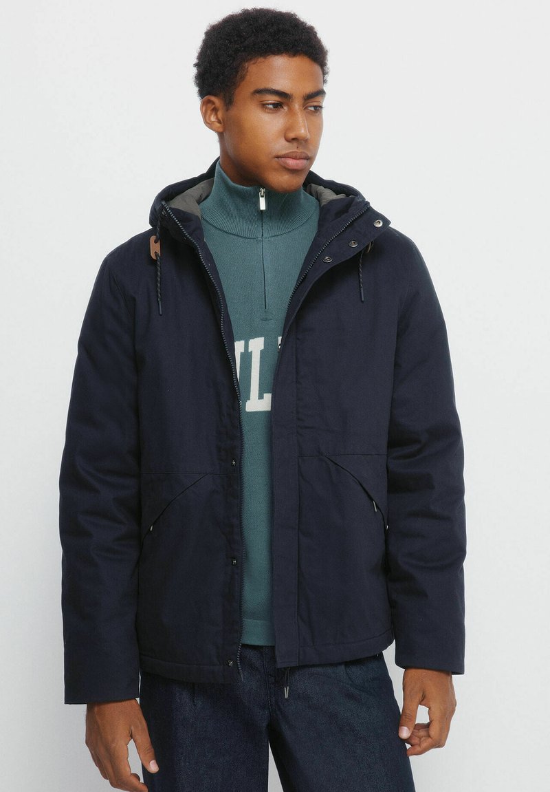 Giacca blu navy con cappuccio, tasche frontali e chiusura con zip. Indossata sopra un dolcevita verde. Il tessuto ha una superficie liscia.