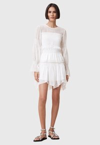 White dress with sheer long sleeves, smocked bodice, layered ruffled hem, and a high-low design. Silver sandals complement the outfit.