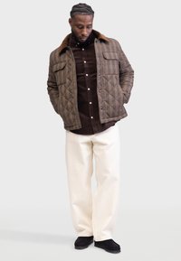 Chaqueta acolchada de cuadros marrones con un cuello de pana marrón, superpuesta sobre una camisa marrón, combinada con pantalones blancos y zapatos negros.