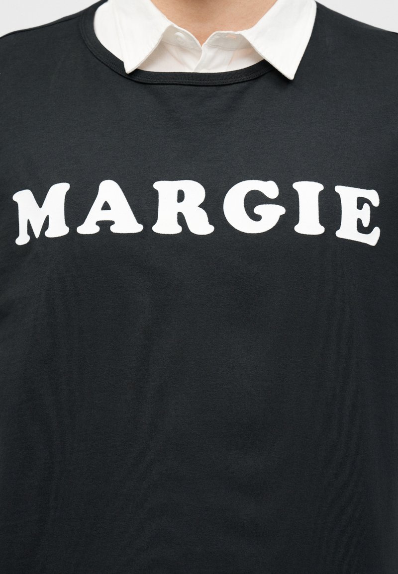 Personne portant un t-shirt noir avec de grandes lettres blanches épelant « MARGIE » sur une chemise blanche à col.