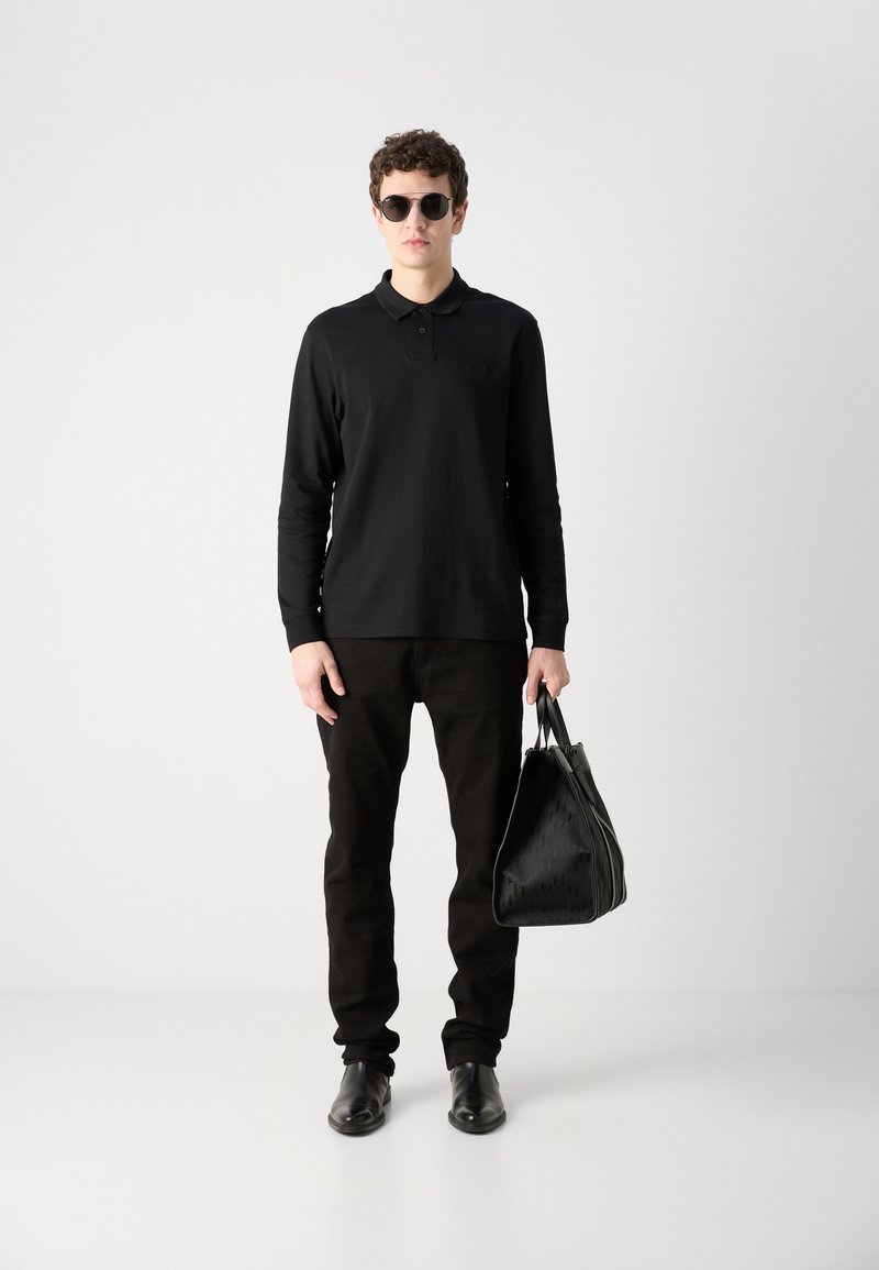 Schwarzes Langarmpolo, schwarze Hose, schwarze Sonnenbrille und schwarze strukturierte Handtasche. Das Modell steht vor einem weißen Hintergrund.