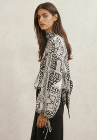 Sort-hvid mønstret bluse med lange ærmer, høj hals og bundne manchetter, med geometriske designs og letvævet stof.