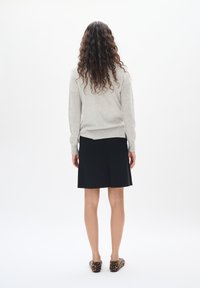 Un pull gris à manches longues, associé à une jupe noire à hauteur du genou et des ballerines à motifs animaliers ; cheveux bouclés visibles. Fond minimaliste.