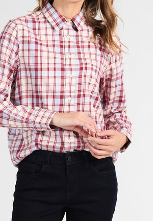 Vrouw draagt een rood-wit geruit overhemd met lange mouwen, ingestopt in zwarte jeans, en past met haar handen één manchetknoop aan.