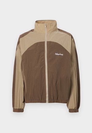 Braune und beige Zip-Jacke mit steigendem Kragen, strukturiertem Stoff, elastischen Bündchen und einem Logo auf der linken Brustseite.
