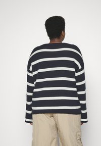 Maglione a righe navy e bianche con vestibilità ampia, maniche lunghe e collo a coste, abbinato a pantaloni beige chiaro con vestibilità rilassata.