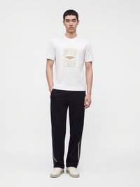 Млад мъж, облечен с бяла тениска Emporio Armani, черни панталони с бели райета и светли маратонки, стои пред обикновен фон.